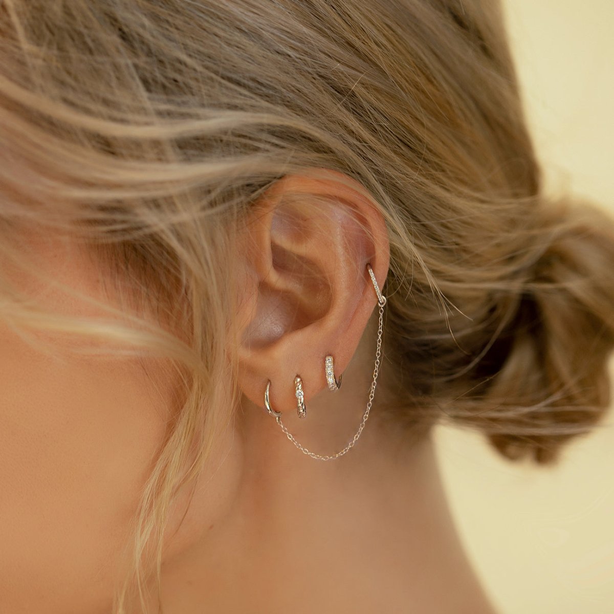 Close-up of woman's ear featuring 14k white gold pave rope huggies, 14k white gold pave huggie single earring, 14k white gold single mini huggie, 14k white gold cartilage pave hoop earring, and 14k white gold chain earring jacket for stud