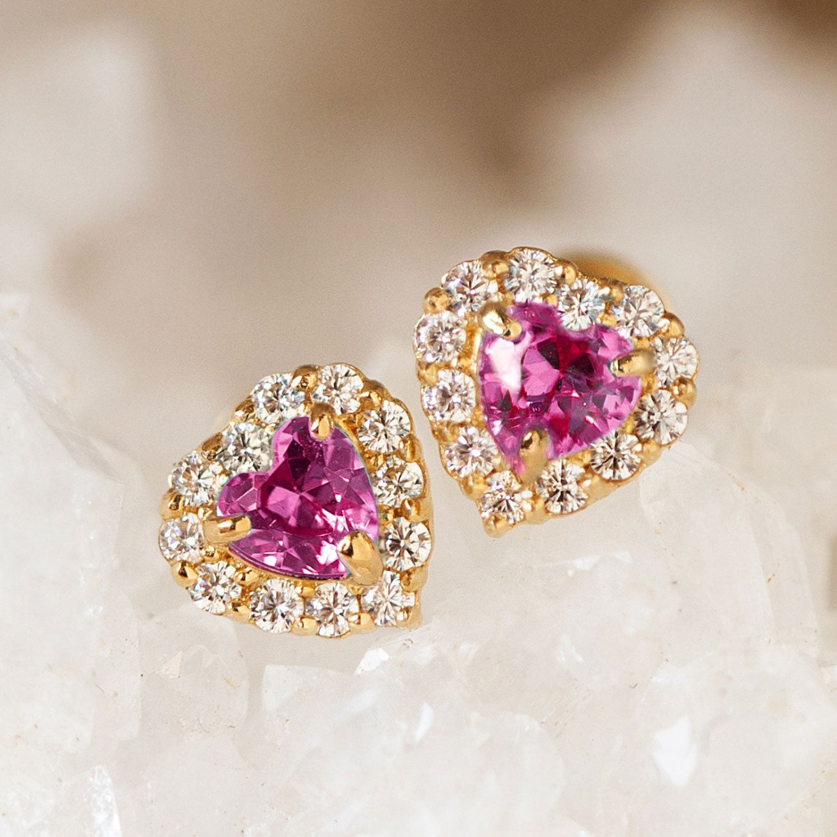 14K gold ruby heart halo studs with pavé-framed pink heart stones, close-up detail on a crystal surface