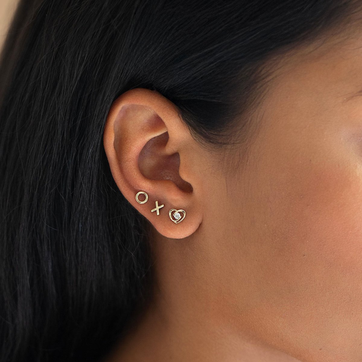 Close-up of an ear wearing 14k gold stud earrings with X and O and a heart stud from AMYO.