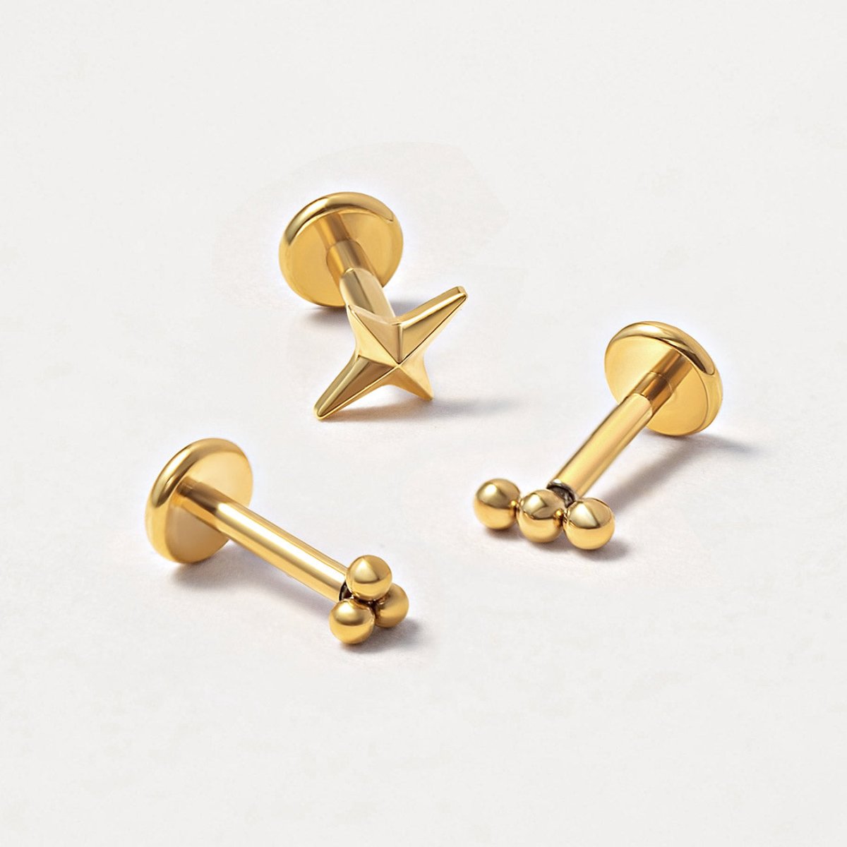 Detailed shot of three gold flat back stud earrings from AMYO featuring Northern Star and beaded styles on a light gray background