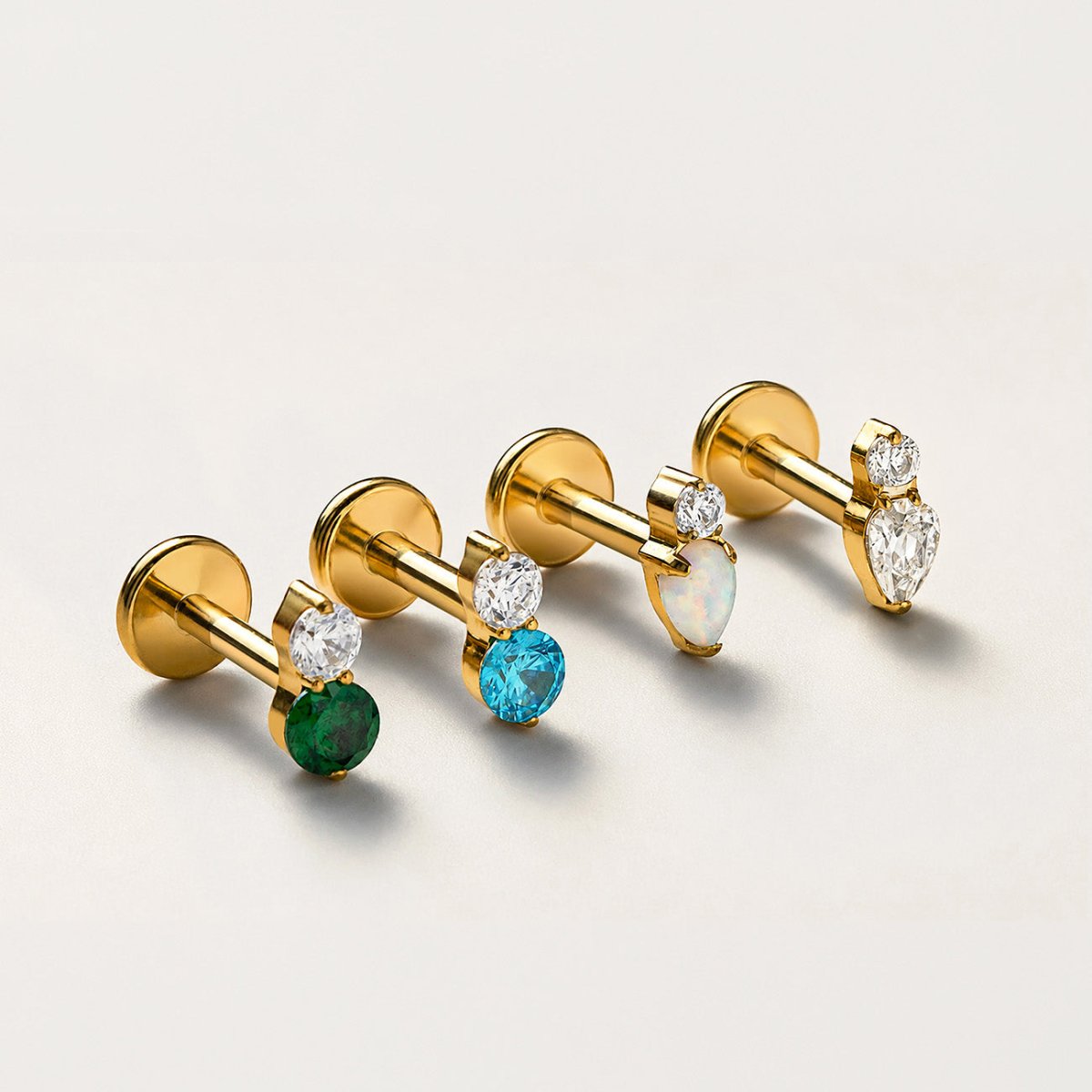 Four gold flat back stud earrings with clear and colored stones on a light beige background | #Gemstone_Emerald