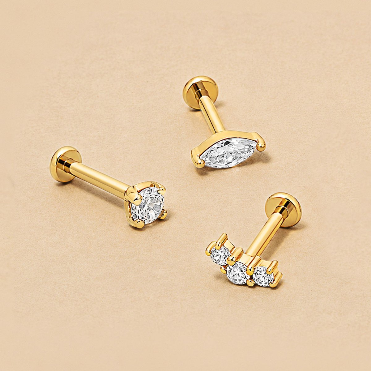 Three 14K gold flat back studs on a beige surface: a round crystal, a marquise crystal and a three-stone curve