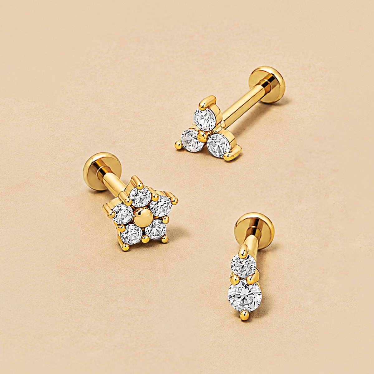 Three 14K gold flat back studs on a beige surface: a clover, a two-crystal duo and a crystal flower