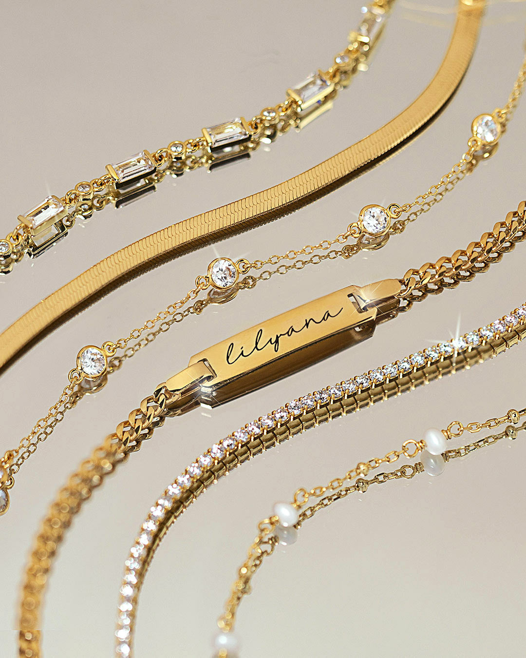 Assorted gold bracelets featuring engraved ID bracelets, crystal tennis bracelets, herringbone chains, and delicate layered styles from AMYO.