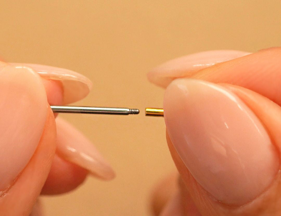 Fingers holding a threaded silver post beside a small gold flatback piece, close-up against a beige background
