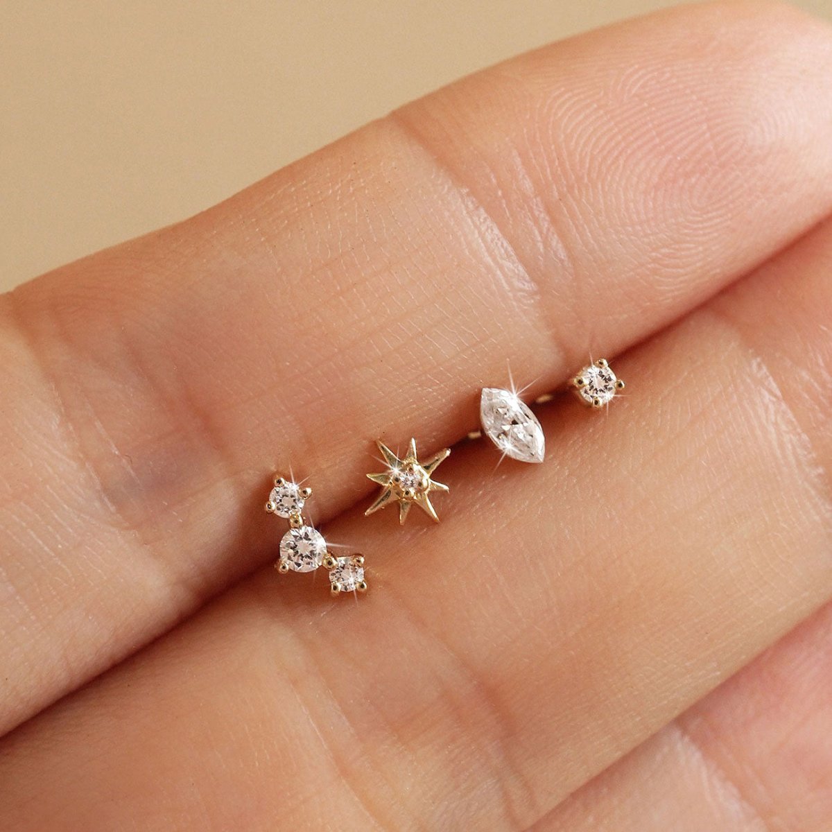 Four tiny 14K gold flat back studs on fingertips: three-stone curve, starburst, marquise and round stud