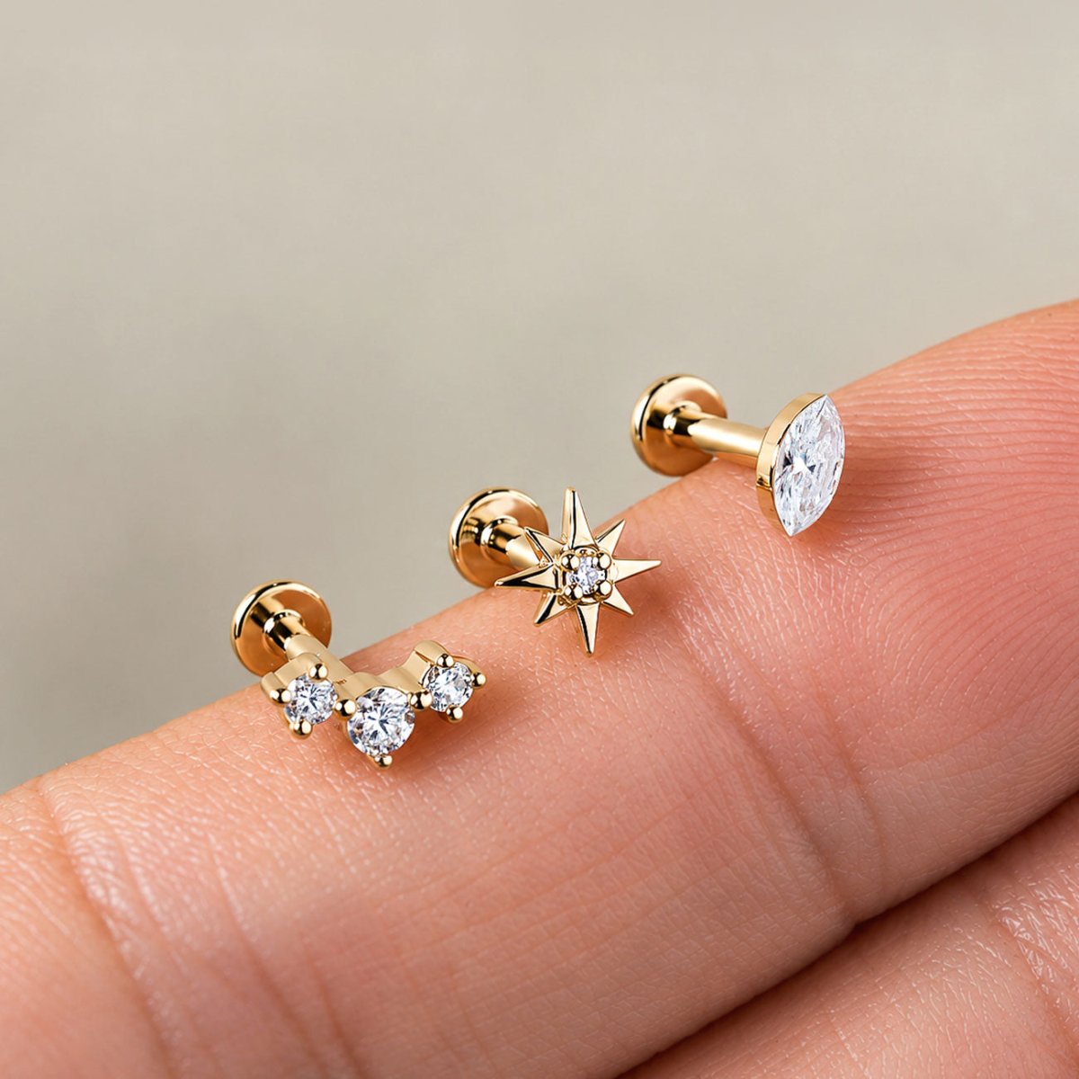 Three 14K gold flat back studs resting on fingertips: a three-stone curve, a star, and a marquise crystal
