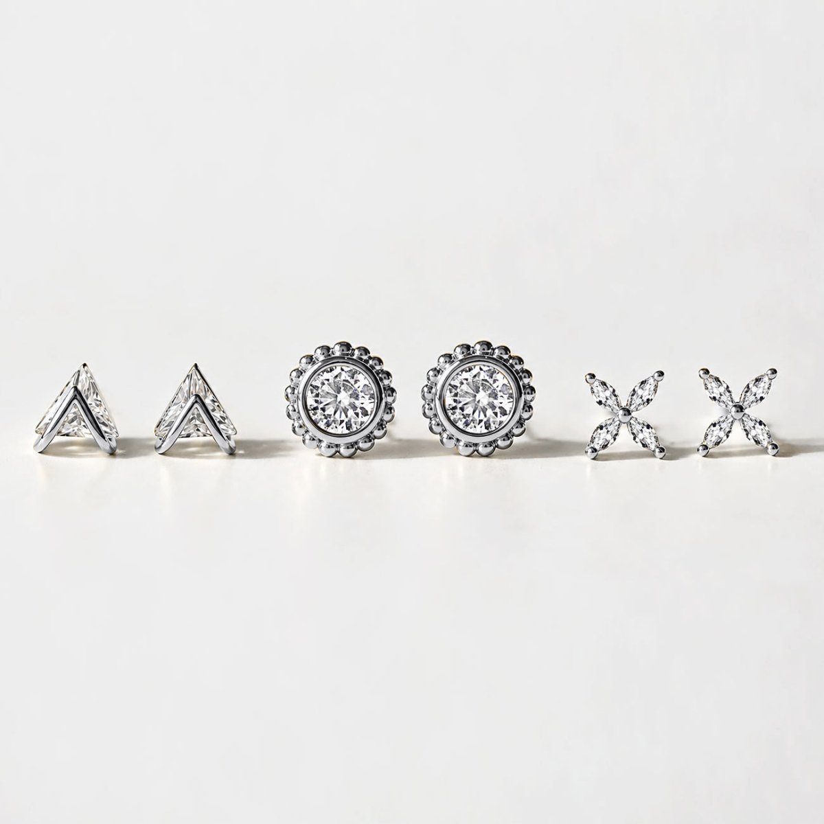 Three pairs of silver studs on a white surface: trinity, beaded halo with round crystal, and four-petal flower