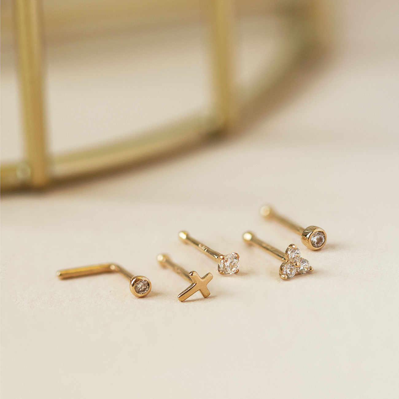 Assorted gold nose studs with clear stones, a cross, and a trio cluster, on cream