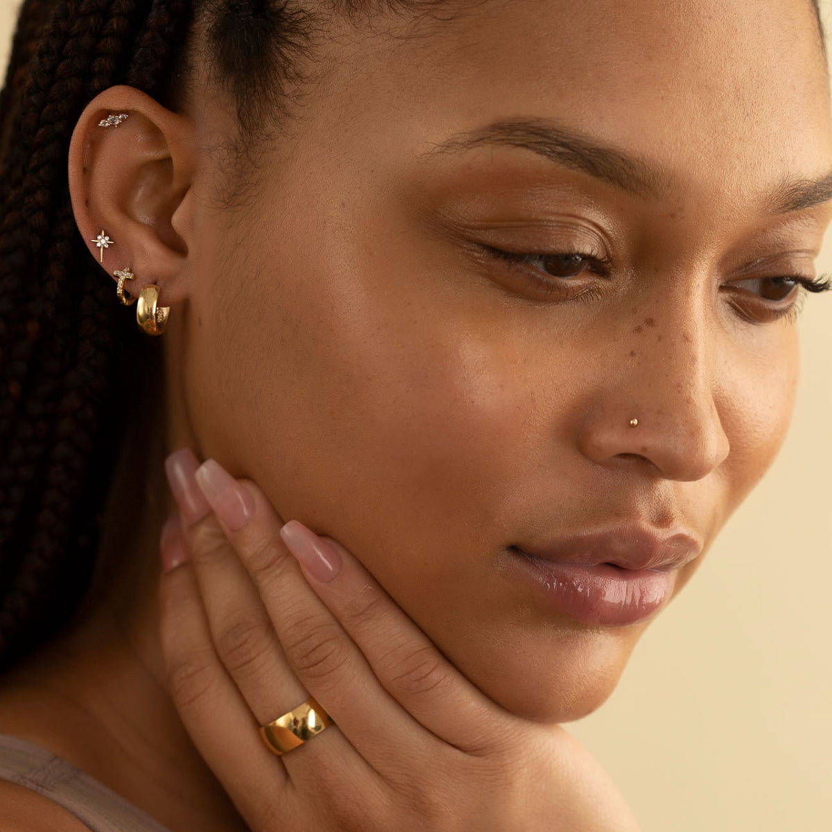 Tiny gold disc nose stud worn on a model's nostril, with stacked ear studs and hoops.
